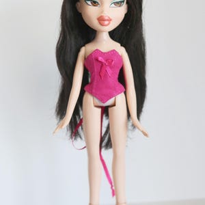 May include: A pink corset with a bow on the front, worn by a doll with long black hair and green eyeshadow. The doll is wearing black high heels.