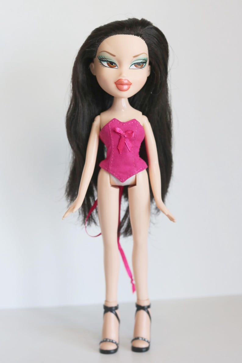 May include: A pink corset with a bow on the front, worn by a doll with long black hair and green eyeshadow. The doll is wearing black high heels.
