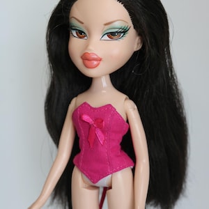 May include: A black-haired doll with green eyeshadow and pink lipstick wearing a pink corset with a bow.