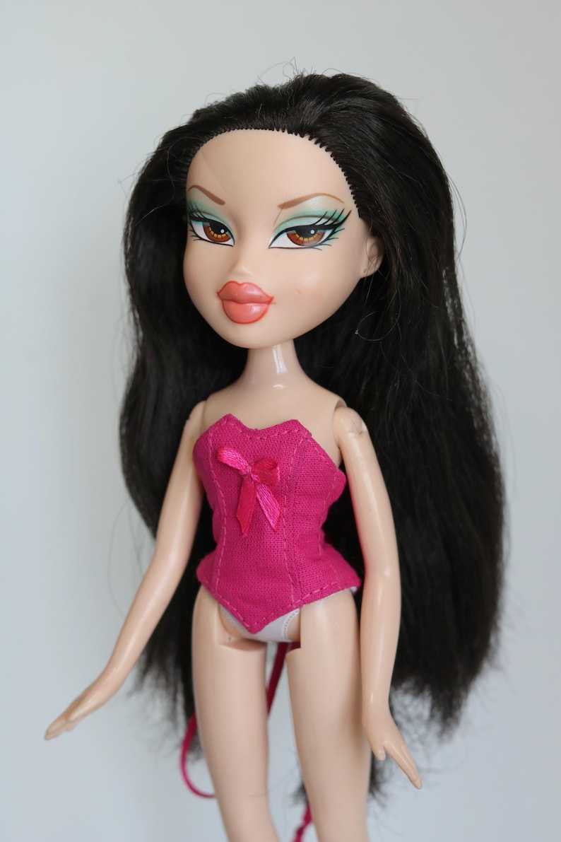May include: A black-haired doll with green eyeshadow and pink lipstick wearing a pink corset with a bow.