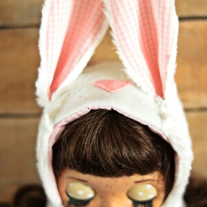 May include: A white bunny hat with pink and white gingham ears. The hat is worn by a doll with brown hair, large eyes, and freckles.