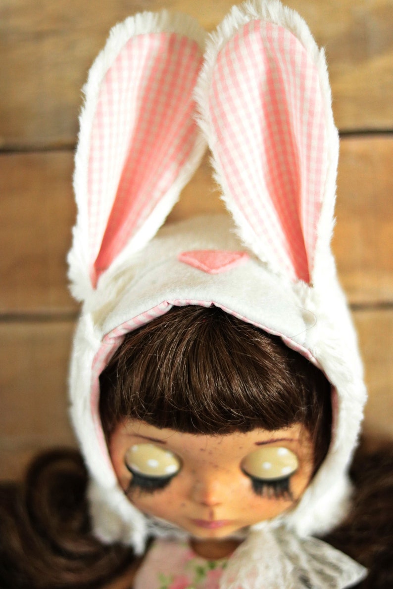 May include: A white bunny hat with pink and white gingham ears. The hat is worn by a doll with brown hair, large eyes, and freckles.