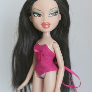 May include: A pink and white doll with long black hair. The doll is wearing a pink corset-style top with a white bow. The doll has green eyeshadow and brown eyes.