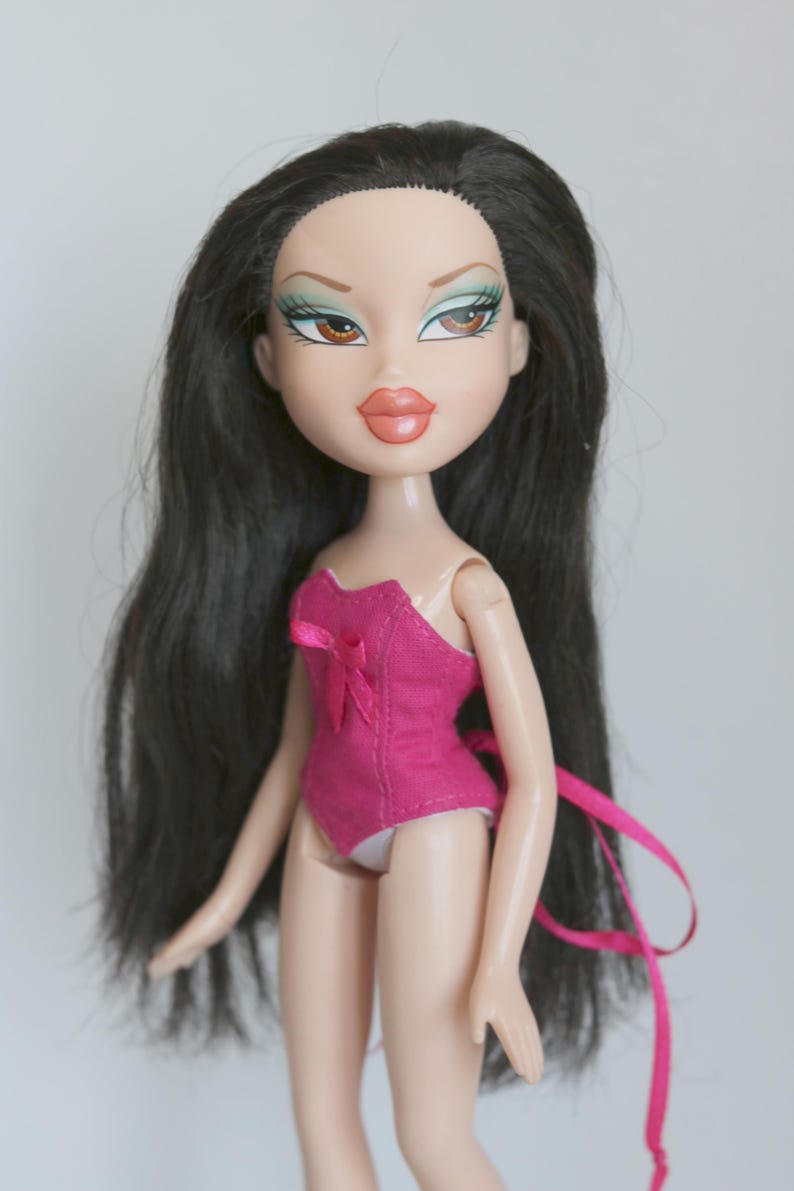 May include: A pink and white doll with long black hair. The doll is wearing a pink corset-style top with a white bow. The doll has green eyeshadow and brown eyes.