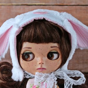 May include: A doll with brown hair wearing a white and pink bunny hat with a white lace collar.