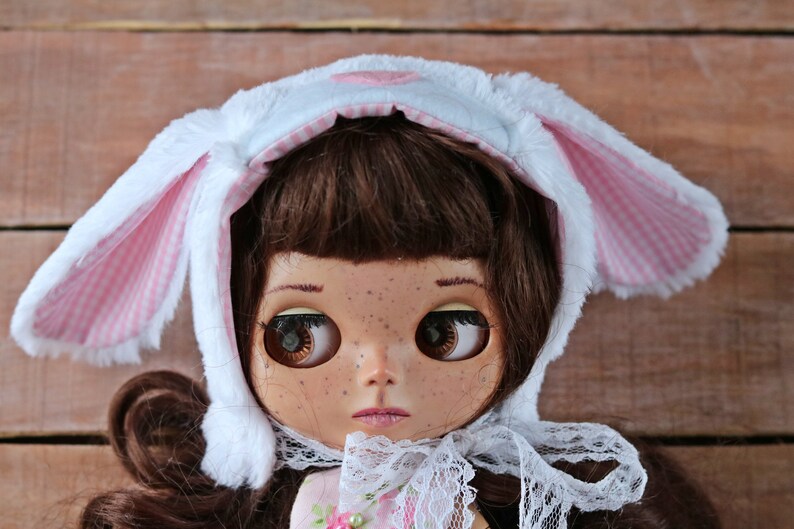 May include: A doll with brown hair wearing a white and pink bunny hat with a white lace collar.