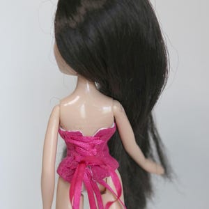 May include: A pink corset with white lace trim and pink ribbon ties. The corset is worn by a doll with long black hair.