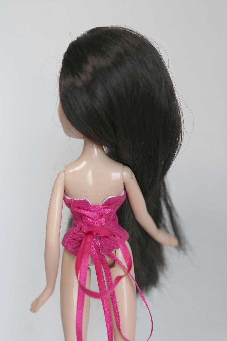 May include: A pink corset with white lace trim and pink ribbon ties. The corset is worn by a doll with long black hair.