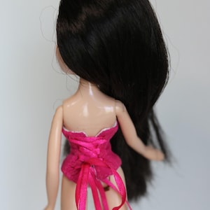 May include: A doll with long black hair wearing a pink corset with white trim and pink ribbon ties.