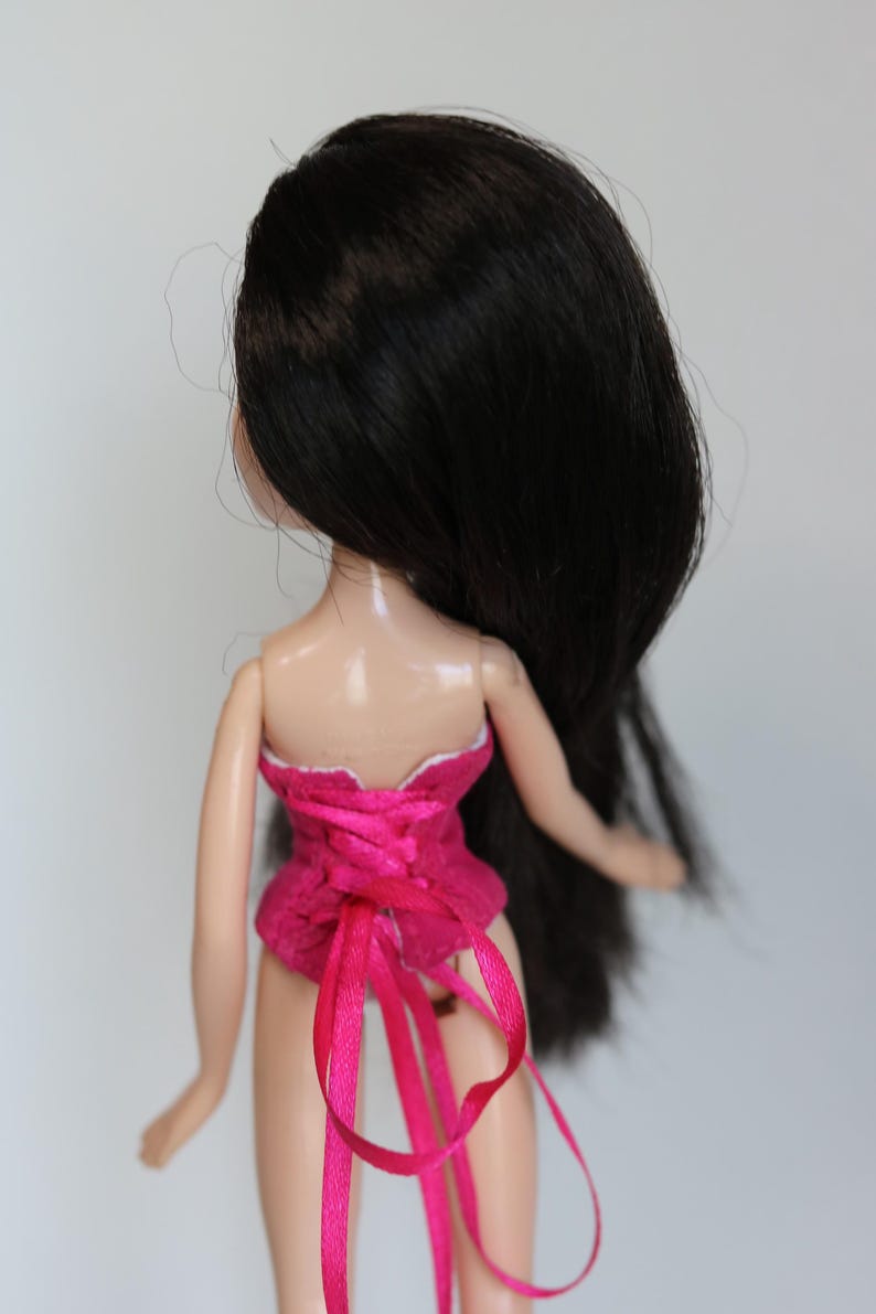 May include: A doll with long black hair wearing a pink corset with white trim and pink ribbon ties.