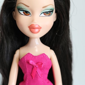 May include: A doll with long black hair, wearing a pink corset with a bow. The doll has green eyeshadow and brown eyes.