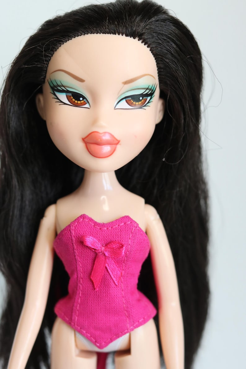 May include: A doll with long black hair, wearing a pink corset with a bow. The doll has green eyeshadow and brown eyes.