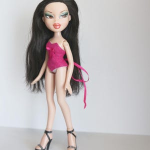 May include: A black-haired doll with green eyeshadow and pink lipstick is wearing a pink swimsuit with a bow and black high-heeled shoes.