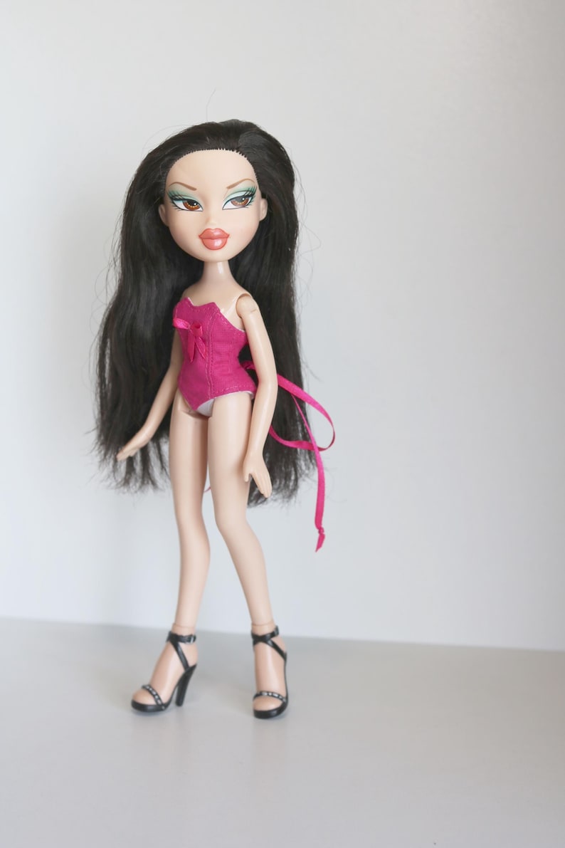 May include: A black-haired doll with green eyeshadow and pink lipstick is wearing a pink swimsuit with a bow and black high-heeled shoes.