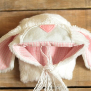May include: White bunny hat with pink and white gingham lining and a pink felt triangle on the top. The hat has a white lace tie.
