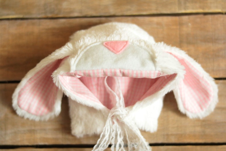 May include: White bunny hat with pink and white gingham lining and a pink felt triangle on the top. The hat has a white lace tie.
