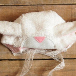 May include: White bunny hat with pink ears and a pink felt nose. The hat has a white lace trim.