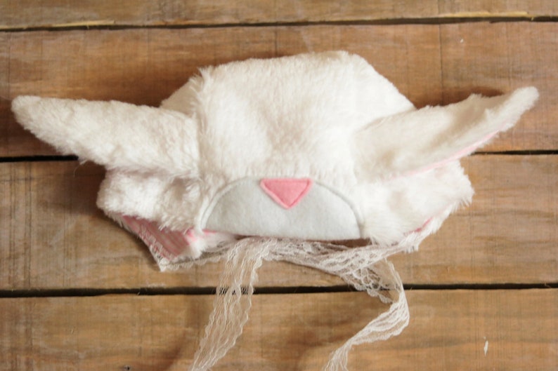 May include: White bunny hat with pink ears and a pink felt nose. The hat has a white lace trim.