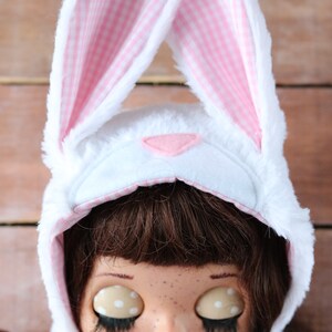 May include: A white bunny hat with pink and white checkered ears. The hat is worn by a doll with brown hair, freckles, and closed eyes. The doll is wearing a white lace collar.