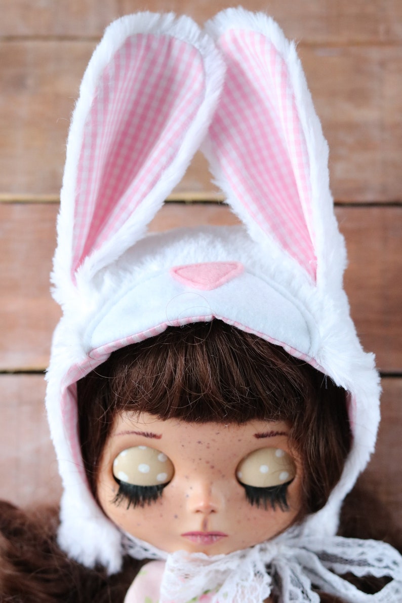 May include: A white bunny hat with pink and white checkered ears. The hat is worn by a doll with brown hair, freckles, and closed eyes. The doll is wearing a white lace collar.
