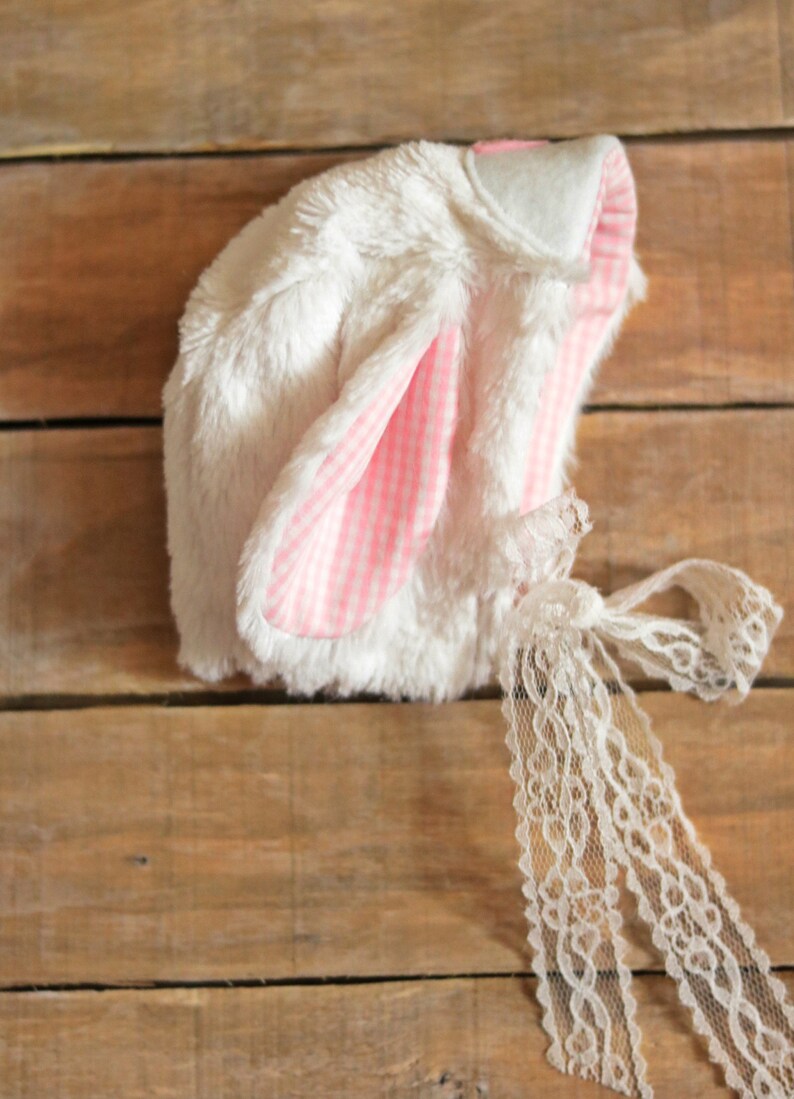 May include: White bunny hat with pink and white gingham ears and a white lace tie.