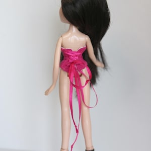 May include: A pink corset with a satin ribbon tied in the back. The doll is wearing black high heels.