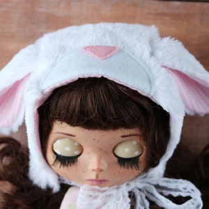 May include: A white plush bunny hat with pink ears and a pink triangle on the front. The hat is worn by a doll with brown hair and large eyes.