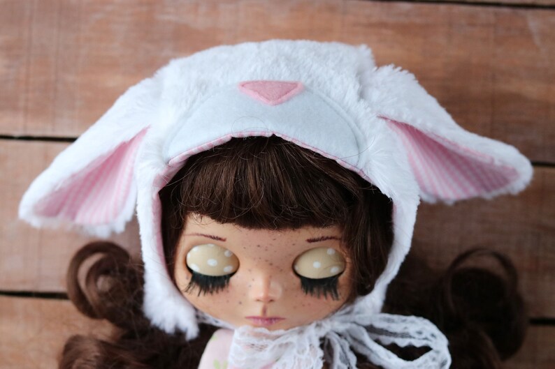 May include: A white plush bunny hat with pink ears and a pink triangle on the front. The hat is worn by a doll with brown hair and large eyes.