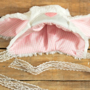 May include: White and pink bunny ear headband with a white lace tie. The headband is made of soft, fluffy fabric and has pink and white gingham lining.