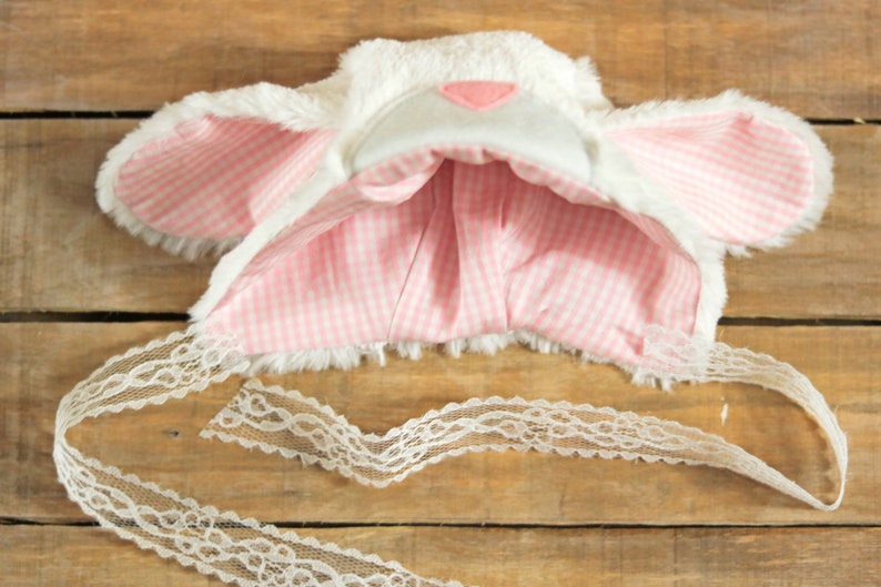 May include: White and pink bunny ear headband with a white lace tie. The headband is made of soft, fluffy fabric and has pink and white gingham lining.