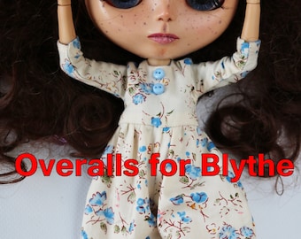 May include: A doll wearing a white and blue floral overall dress with blue buttons. The doll has long brown hair and blue eyes. The text "Overalls for Blythe" is written in red letters below the doll.