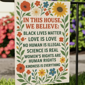 May include: A rectangular garden flag with the text "IN THIS HOUSE, WE BELIEVE: BLACK LIVES MATTER LOVE IS LOVE NO HUMAN IS ILLEGAL SCIENCE IS REAL WOMEN'S RIGHTS ARE HUMAN RIGHTS KINDNESS IS EVERYTHING." The flag is decorated with colorful flowers.