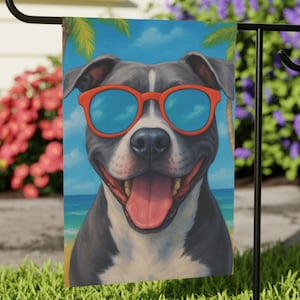 May include: A garden flag featuring a smiling pit bull wearing orange sunglasses with blue lenses. The dog is depicted on a beach scene with palm trees and a blue sky. The flag is displayed on a black metal stand, set in a grassy yard.