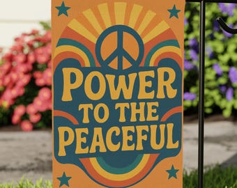 May include: A garden flag with the words "POWER TO THE PEACEFUL" in a retro design. The flag features a peace symbol, a rainbow, and stars on an orange background. The flag is displayed on a black metal stand in a grassy yard.