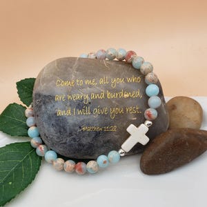May include: A beaded bracelet with light blue, peach, and brown beads, featuring a white cross charm. The bracelet is draped over a gray stone with the text "Come to me, all you who are weary and burdened, and I will give you rest. Matthew 11:28."