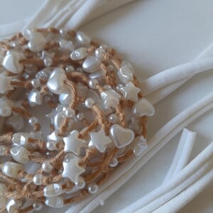 Puede incluir: Un accesorio hecho a mano con una base marrón tejida adornada con cuentas de perlas blancas, cuentas en forma de estrella y cuentas en forma de corazón. Tiras de tela blanca se extienden desde el accesorio, creando un diseño delicado.