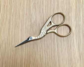 May include: Gold-coloured scissors with a bird-shaped design. The scissors have a small, round hole in the beak of the bird.