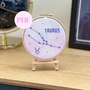 May include: A white embroidery hoop with a purple cross-stitch design of the Taurus constellation. The hoop is on a wooden stand and the text "Taurus" is stitched in purple.