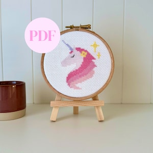 Puede incluir: Un patrón de punto de cruz de un unicornio rosa y blanco en un aro de bordado de madera sobre un caballete de madera. El unicornio tiene un cuerno morado y una flor amarilla en la cabeza. Hay estrellas amarillas en el fondo.