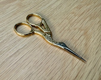 May include: Gold-coloured scissors with a bird-shaped handle. The scissors have two rings and a pointed tip.