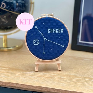 May include: A cross-stitch kit featuring the Cancer constellation on a navy blue background. The design includes the word "CANCER" and the Cancer zodiac symbol in white thread, framed in a wooden hoop and displayed on a small wooden easel.
