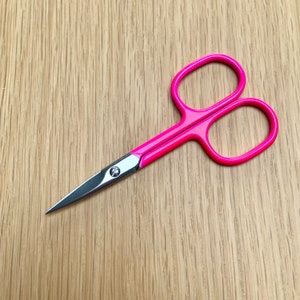 May include: A pair of pink nail clippers with a silver blade. The clippers are on a wooden surface.