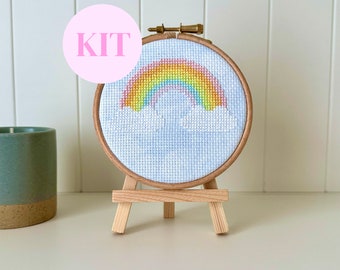 May include: A cross-stitch kit featuring a rainbow design, displayed in a wooden embroidery hoop. The rainbow is composed of various colors, set against a blue sky with white clouds. The word "KIT" is printed on a pink circle.