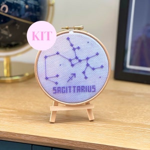 May include: A cross-stitch kit featuring the Sagittarius constellation in purple thread on a light blue fabric. The design is displayed in a wooden embroidery hoop and mounted on a small wooden easel. The word "SAGITTARIUS" is stitched below the constellation.