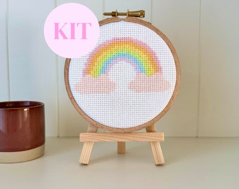 May include: A cross-stitch kit featuring a rainbow design, displayed in a wooden hoop and set on a small wooden easel. The rainbow is composed of various colors, including yellow, orange, pink, blue, and green. The word "KIT" is in a pink circle.