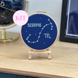 May include: A cross-stitch kit featuring a navy blue fabric with the word "SCORPIO" and the constellation design in white thread. The kit is displayed in a wooden embroidery hoop and sits on a small wooden easel.