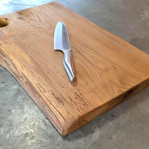 Può includere: Un tagliere rettangolare in legno con bordo naturale e un foro nell'angolo in alto a sinistra. Un coltello in acciaio inossidabile con manico strutturato è appoggiato sulla tavola. Il legno è marrone chiaro.