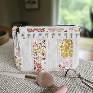 May include: A rectangular zippered pouch featuring a patchwork design of cream fabric with white polka dots and floral patterns in mustard yellow, rust orange, and soft pink. A white fabric ruler strip is sewn across the top, measuring 6 inches.