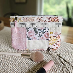 May include: A rectangular quilted zipper pouch featuring a patchwork design of floral and botanical patterns in shades of pink, cream, and mustard yellow. A printed ruler graphic runs across the top edge. A light pink ribbon is attached to the zipper pull.