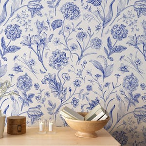 May include: A wallpaper design with a repeating pattern of blue floral illustrations on a white backdrop. The design showcases a variety of flowers and leaves, offering a traditional and refined look. A wooden chest of drawers is in the foreground, with a vase, box, candles, and a bowl.